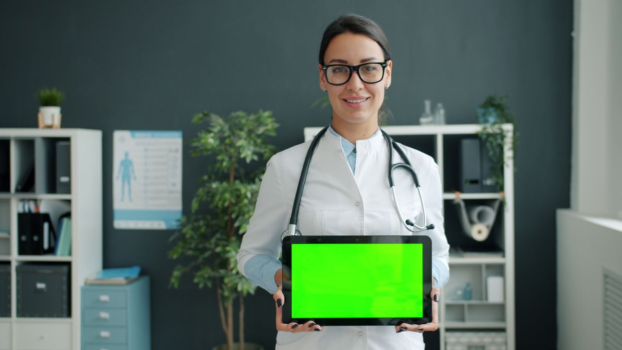 about-us Female doctor smiling and holding a tablet with green screen in a modern office.
