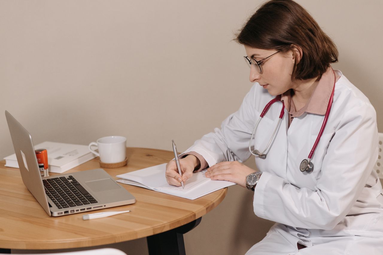 services-03 Woman doctor with stethoscope writing at desk with laptop and notes.